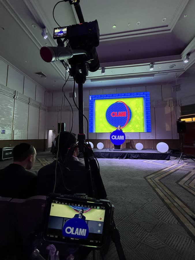 The camera on a tripod in the Olami ballroom, the speaker at the podium showing on the camera's screen